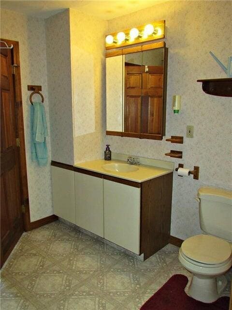 half bathroom with vanity, baseboards, wallpapered walls, and toilet