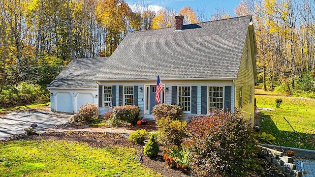 cape cod home with a front yard, an attached garage, a chimney, a shingled roof, and driveway