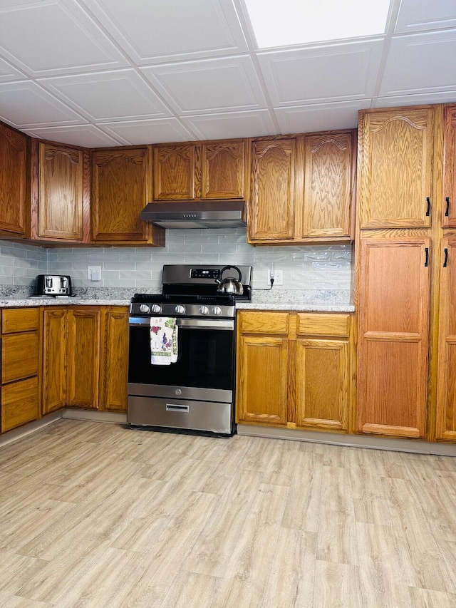 kitchen with light wood finished floors, decorative backsplash, light stone countertops, under cabinet range hood, and stainless steel range with gas stovetop