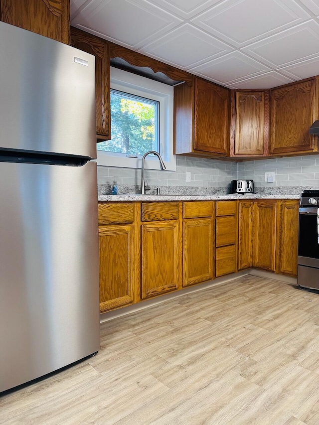 kitchen featuring light stone countertops, light wood-style floors, appliances with stainless steel finishes, backsplash, and brown cabinets