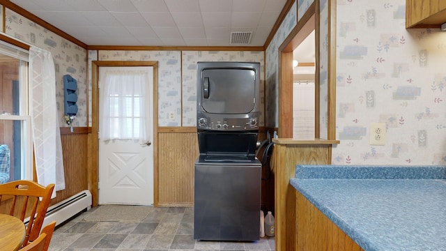 laundry area with ornamental molding, baseboard heating, stacked washer and clothes dryer, and wooden walls