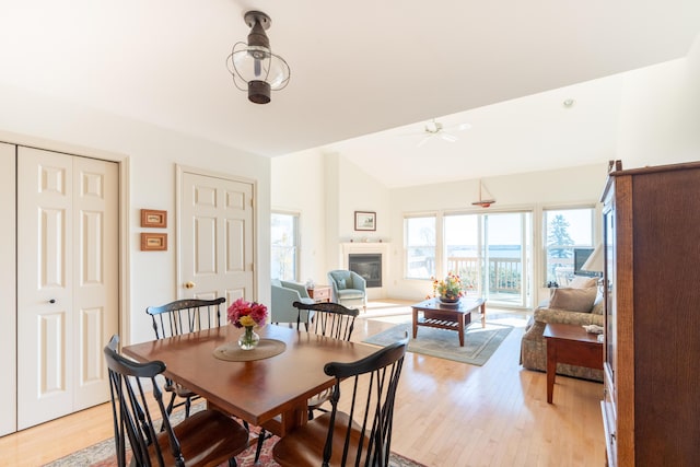 dining room with a ceiling fan, light wood finished floors, a glass covered fireplace, and vaulted ceiling