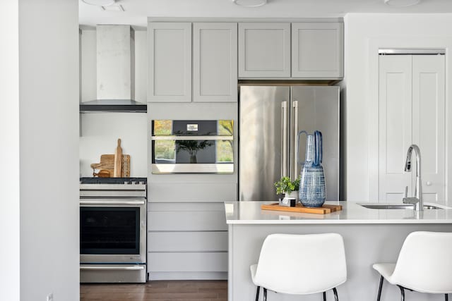 kitchen featuring wall chimney range hood, dark wood-type flooring, stainless steel appliances, sink, and a breakfast bar