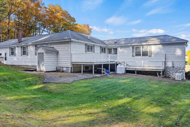 rear view of house featuring roof with shingles, a wooden deck, a chimney, a lawn, and a patio