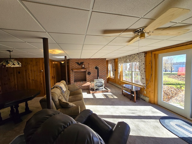 carpeted living room featuring a paneled ceiling, a baseboard radiator, ceiling fan, a wood stove, and wooden walls