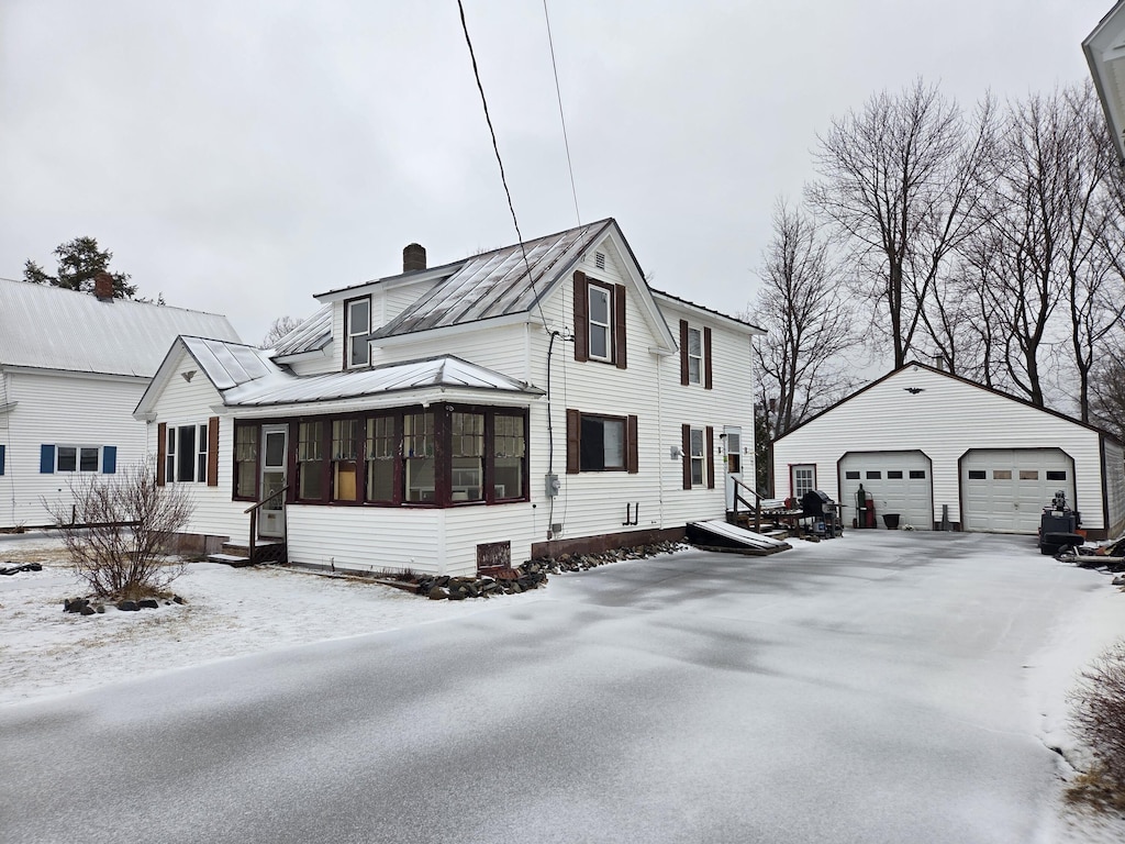 snow covered property featuring a chimney, an outdoor structure, metal roof, a sunroom, and a detached garage