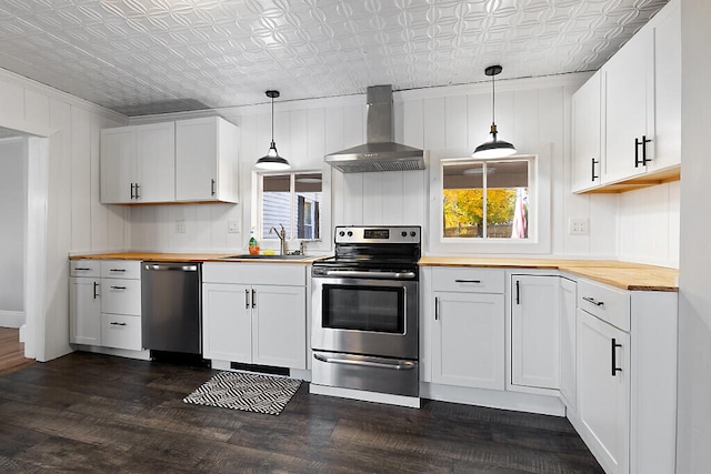 kitchen featuring wall chimney range hood, sink, appliances with stainless steel finishes, white cabinetry, and decorative light fixtures