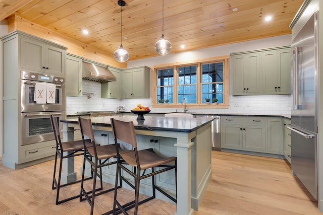 kitchen featuring wall chimney range hood, hanging light fixtures, a kitchen island, light wood-type flooring, and stainless steel appliances