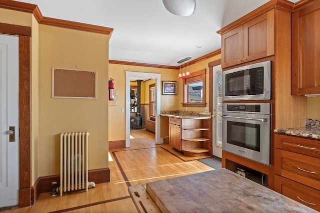 kitchen with radiator heating unit, light stone counters, pendant lighting, light wood-type flooring, and appliances with stainless steel finishes