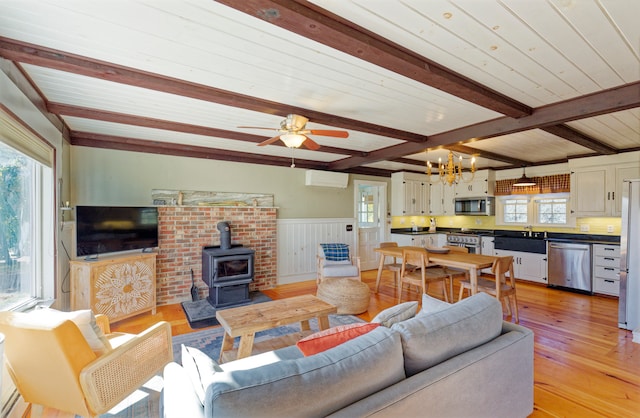 living room featuring light hardwood / wood-style floors, ceiling fan with notable chandelier, beam ceiling, a wood stove, and an AC wall unit