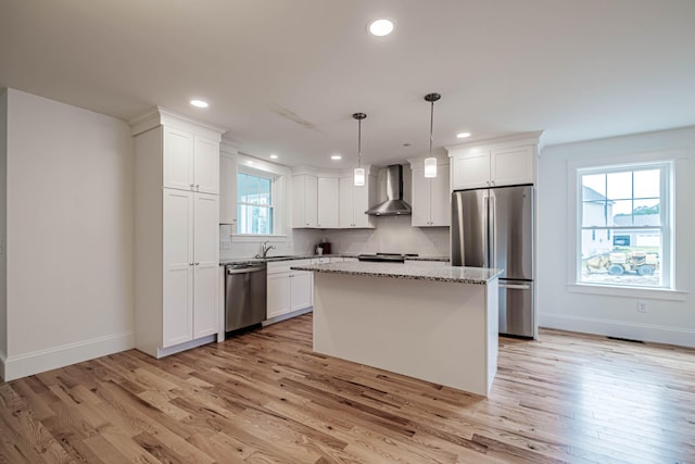 kitchen featuring appliances with stainless steel finishes, wall chimney range hood, light hardwood / wood-style flooring, a center island, and hanging light fixtures