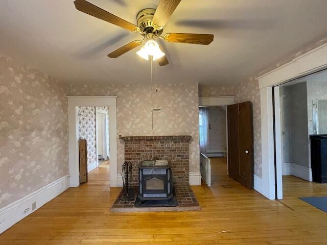 unfurnished living room featuring light wood-type flooring, a wood stove, ceiling fan, and a baseboard heating unit