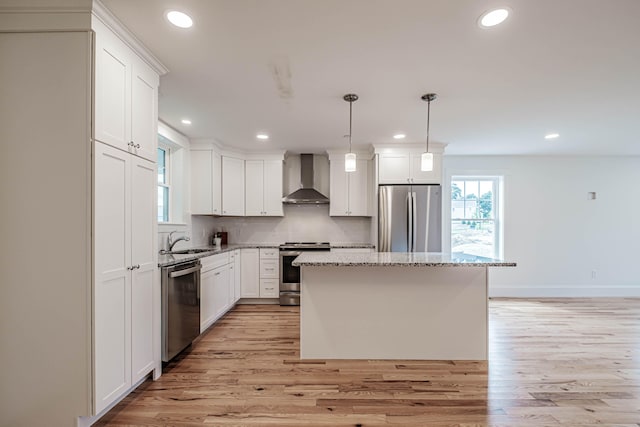 kitchen with appliances with stainless steel finishes, wall chimney exhaust hood, decorative light fixtures, a center island, and light hardwood / wood-style floors