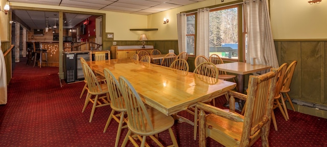 dining room with a paneled ceiling, wooden walls, and dark colored carpet