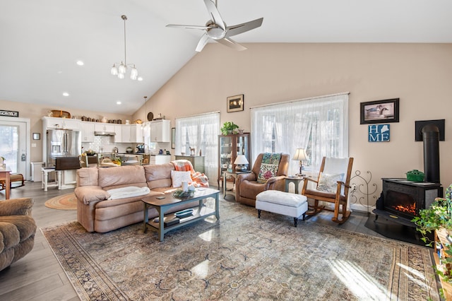 living room with hardwood / wood-style flooring, ceiling fan with notable chandelier, a wood stove, and high vaulted ceiling