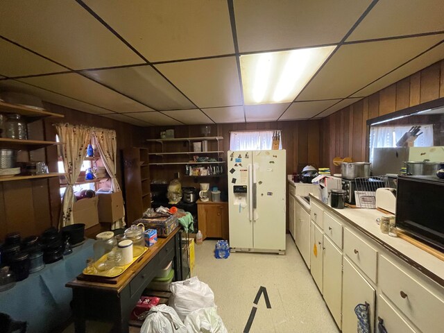 kitchen with a drop ceiling, white fridge with ice dispenser, and wooden walls