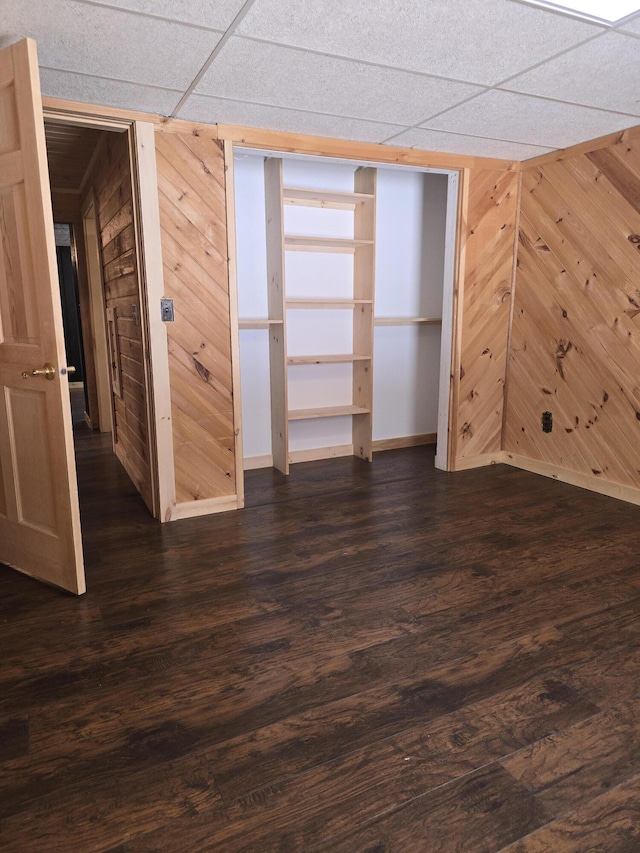 unfurnished bedroom featuring a drop ceiling, baseboards, dark wood-style flooring, and wood walls