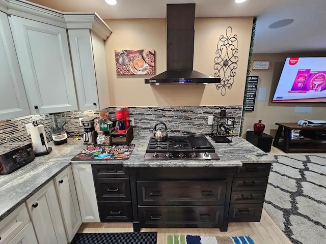 kitchen with backsplash, wall chimney range hood, gas stovetop, light stone countertops, and white cabinetry