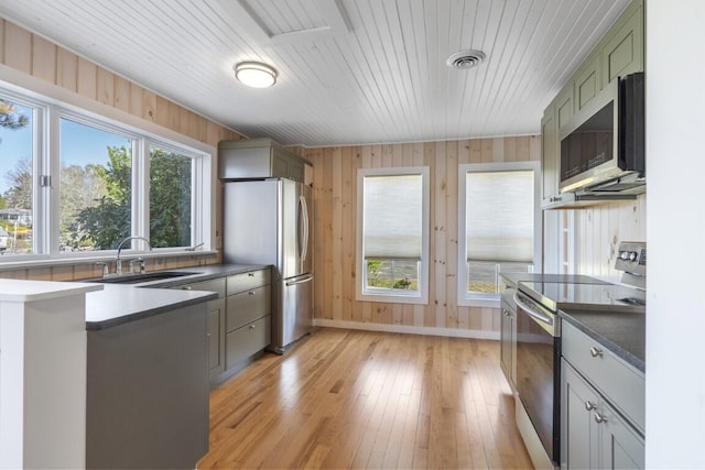 kitchen with light wood-type flooring, stainless steel appliances, sink, green cabinetry, and wood walls