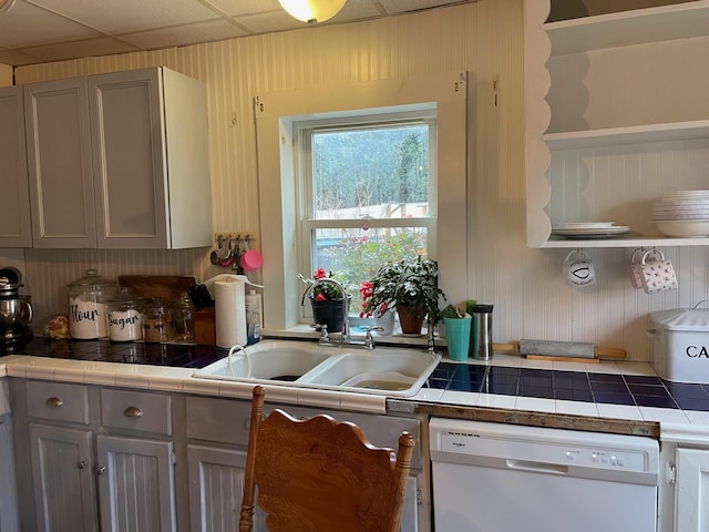 kitchen with a paneled ceiling, tile countertops, white dishwasher, and gray cabinetry