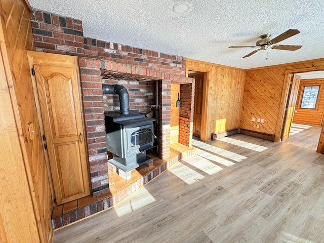 unfurnished living room featuring a textured ceiling, ceiling fan, wooden walls, light hardwood / wood-style flooring, and a wood stove