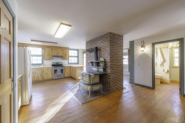 kitchen featuring stainless steel range with gas stovetop, a wood stove, light brown cabinets, white refrigerator, and light hardwood / wood-style flooring