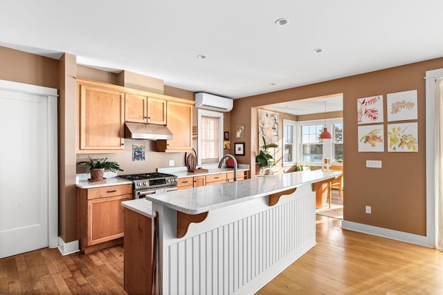 kitchen with gas range, light stone counters, a center island with sink, a wall unit AC, and light hardwood / wood-style floors