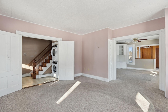 carpeted empty room with a baseboard heating unit, baseboards, stairway, stacked washer and dryer, and a ceiling fan