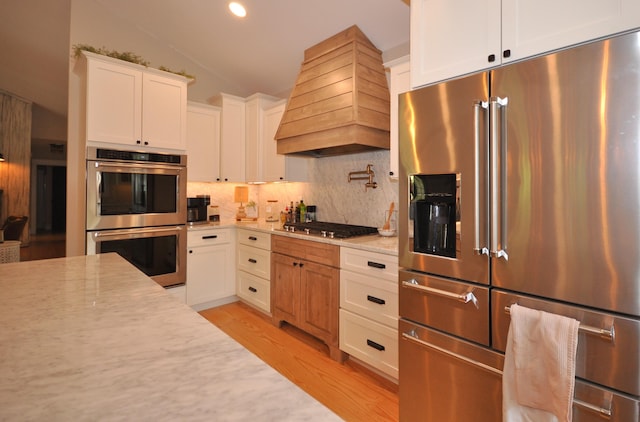 kitchen with appliances with stainless steel finishes, light wood-type flooring, premium range hood, light stone counters, and white cabinetry