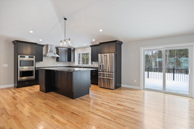 kitchen with pendant lighting, light wood-style flooring, appliances with stainless steel finishes, a kitchen island, and wall chimney range hood