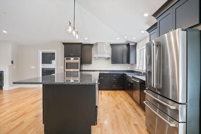 kitchen featuring a center island, light wood finished floors, stainless steel appliances, dark stone countertops, and wall chimney exhaust hood