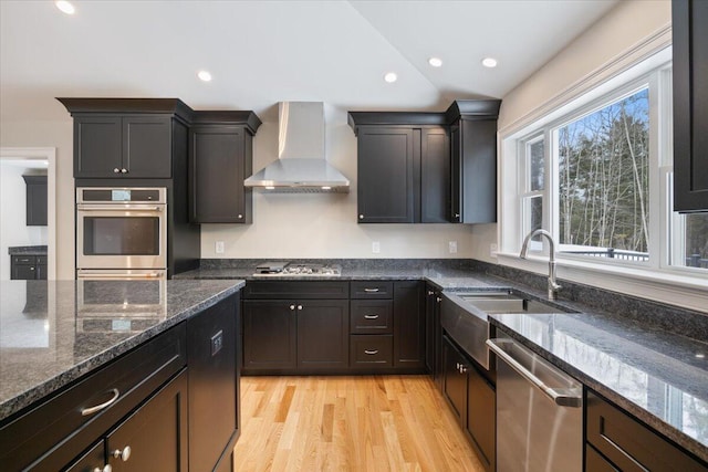 kitchen featuring dark stone counters, stainless steel appliances, wall chimney range hood, and a sink