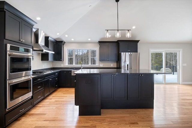 kitchen featuring light wood-style flooring, appliances with stainless steel finishes, a kitchen island, wall chimney range hood, and dark stone countertops