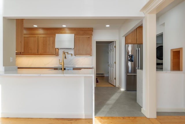 kitchen with decorative backsplash, stainless steel refrigerator with ice dispenser, kitchen peninsula, a baseboard heating unit, and stacked washer / drying machine