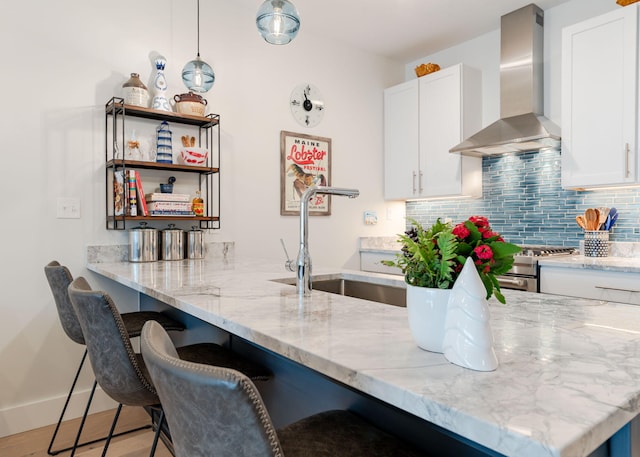 kitchen featuring white cabinetry, sink, hanging light fixtures, wall chimney range hood, and light stone counters