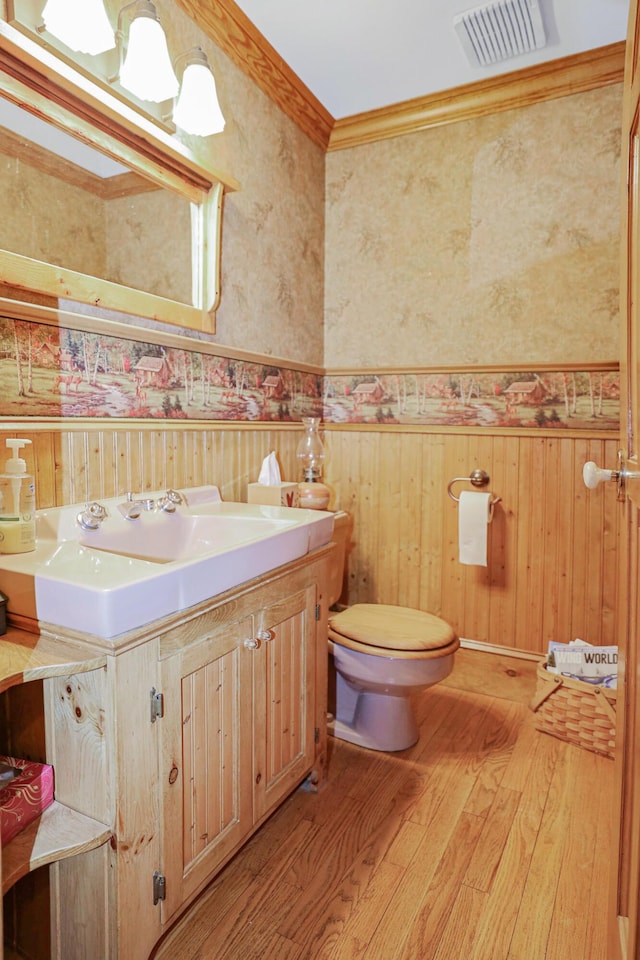 half bath featuring crown molding, visible vents, wainscoting, wood finished floors, and wallpapered walls