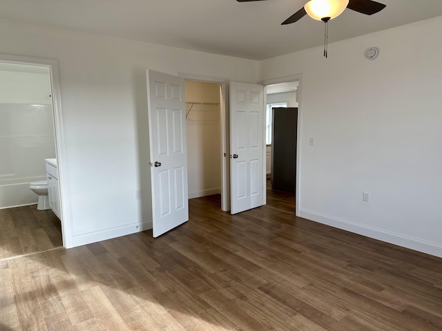 unfurnished bedroom with ceiling fan, a walk in closet, ensuite bathroom, and dark wood-type flooring