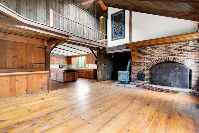 unfurnished living room with a wood stove, high vaulted ceiling, wooden walls, ceiling fan, and light wood-type flooring