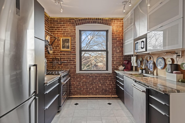 kitchen with sink, brick wall, dark stone counters, light tile patterned flooring, and appliances with stainless steel finishes