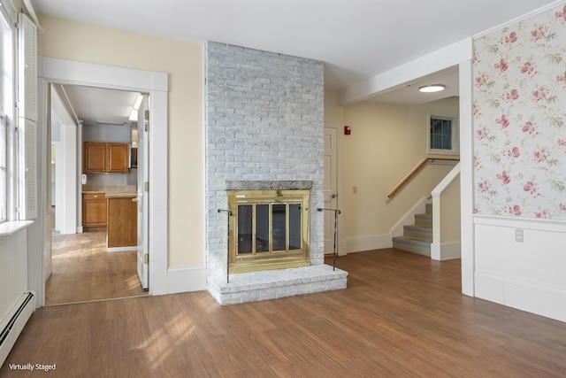 unfurnished living room featuring a wainscoted wall, stairway, baseboard heating, a brick fireplace, and wood finished floors