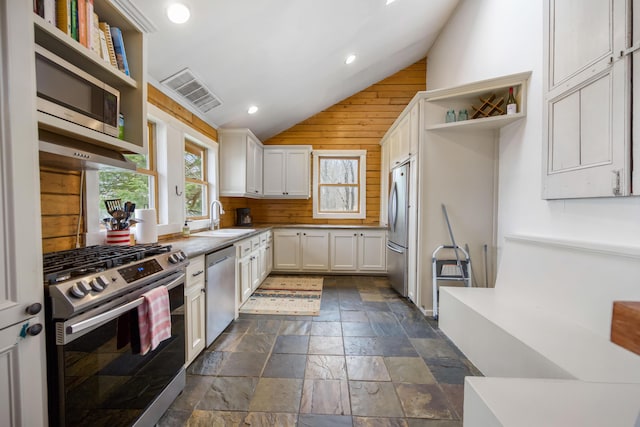 kitchen featuring stainless steel appliances, sink, white cabinetry, lofted ceiling, and wood walls