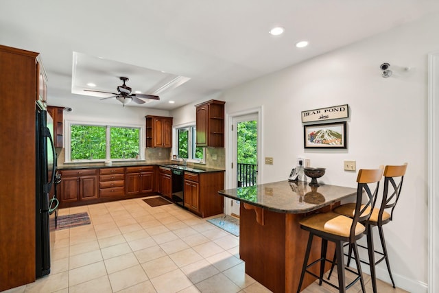 kitchen with a kitchen breakfast bar, a raised ceiling, black appliances, kitchen peninsula, and dark stone counters