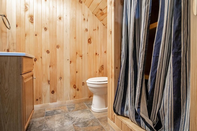 full bath with a shower with shower curtain, baseboards, stone finish floor, wood walls, and toilet