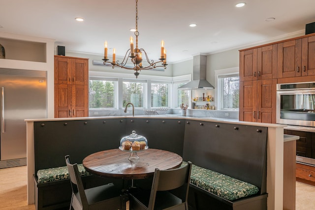 kitchen featuring stainless steel appliances, wall chimney exhaust hood, a chandelier, brown cabinetry, and crown molding