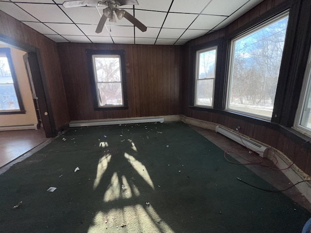 empty room with ceiling fan, a baseboard radiator, a drop ceiling, and wooden walls