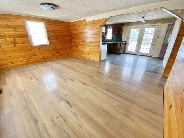 unfurnished living room featuring french doors, wooden walls, a textured ceiling, and light hardwood / wood-style floors