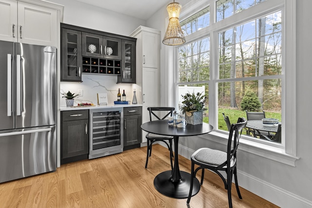 bar featuring backsplash, pendant lighting, stainless steel refrigerator, light wood-type flooring, and beverage cooler