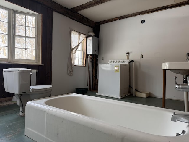 bathroom featuring water heater, beam ceiling, a tub to relax in, washer / clothes dryer, and toilet