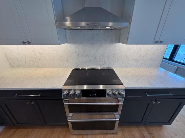 kitchen with wall chimney range hood, backsplash, light stone counters, high end stainless steel range, and light hardwood / wood-style floors