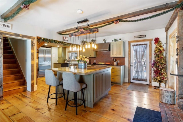 kitchen featuring wall chimney range hood, a center island, stainless steel built in refrigerator, beamed ceiling, and dark countertops